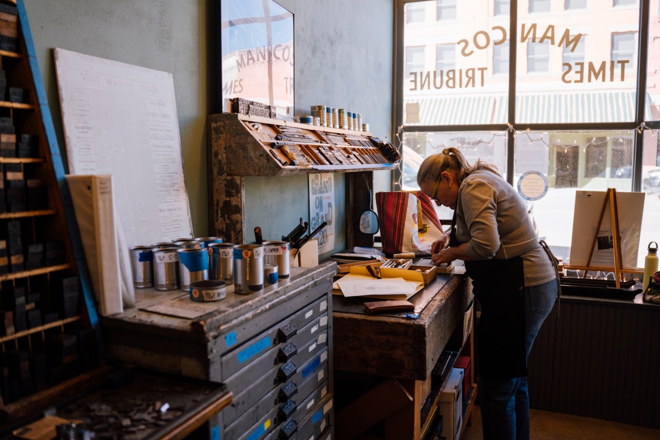 A woman wearing an apron and glasses stands over a workbench, beside a window at Mancos Common Press. On a counter new her sit cans of paint with different labels on them. 